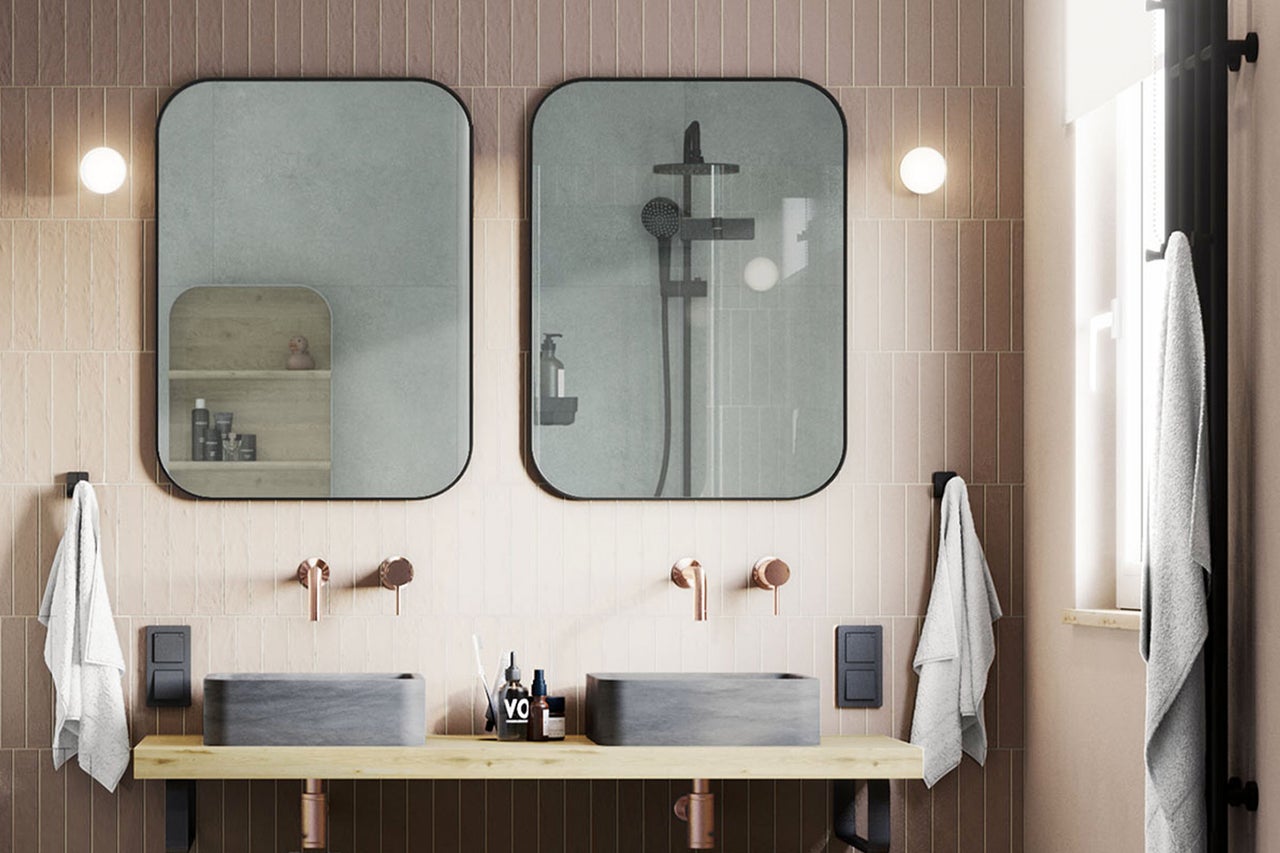 Modernes Badezimmer mit zwei grauen Stein-Aufsatzwaschbecken auf Holzplatte, Kupferarmaturen und zwei abgerundeten Spiegeln vor schmalen Wandfliesen.