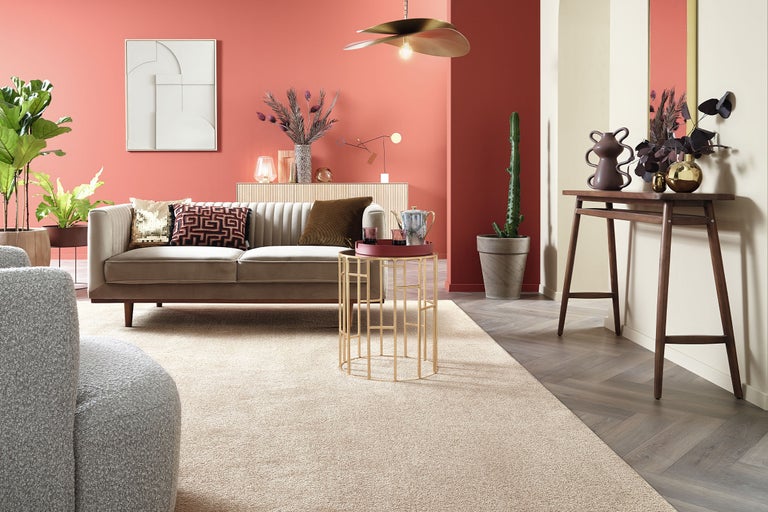 Wohnzimmer mit beigem Samtsofa, goldenem Beistelltisch, hellem Teppich und dunklem Konsolentisch vor korallenroter Wand.