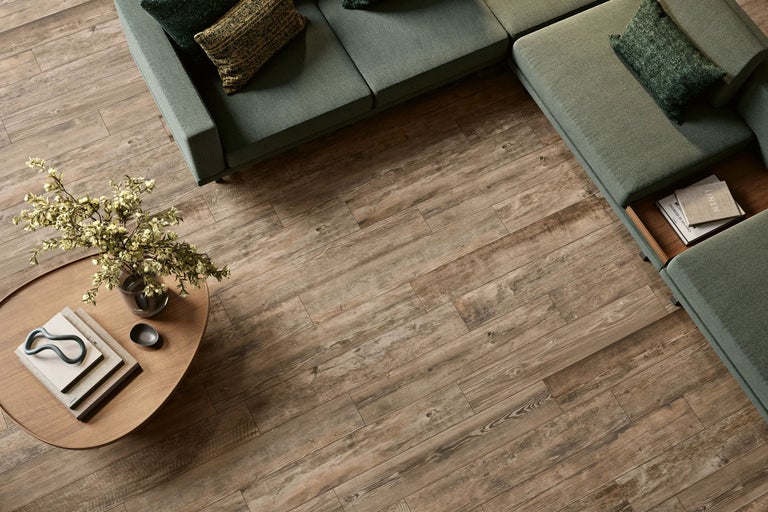 Wohnzimmer mit Bodenbelag in Holzoptik, grünem Ecksofa und rundem Holztisch in einer hellen Atmosphäre.