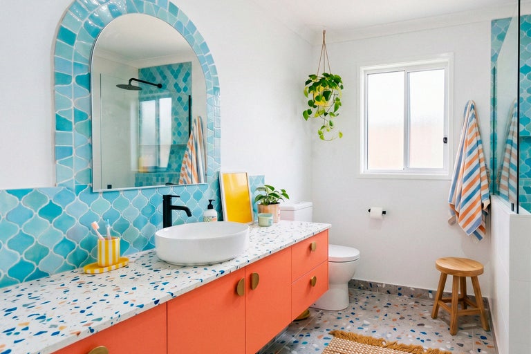 Modernes Badezimmer mit orangem Waschtisch, Terrazzo-Arbeitsplatte, Aufsatzwaschbecken, blauen Mosaikfliesen, Rundbogenspiegel und Holzschemel.