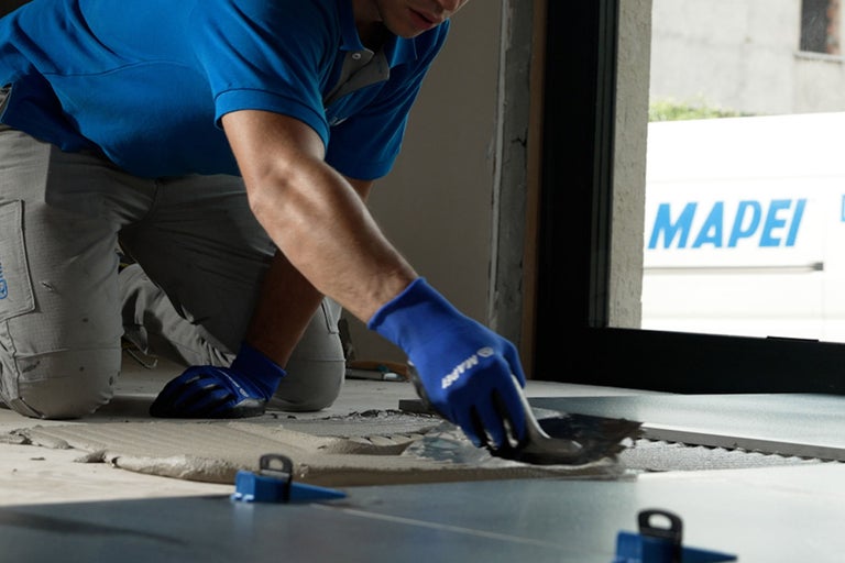 Handwerker trägt Fliesenkleber mit einer Zahnkelle auf, Nivelliersystem im Vordergrund und Mapei Logo im Hintergrund.