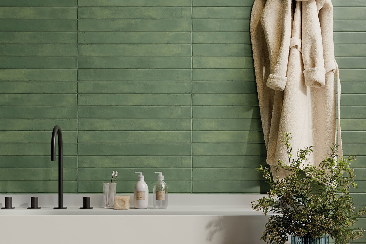 Helles Badezimmer mit grünen horizontalen Wandfliesen, schwarzen Armaturen, weißer Ablage mit Seifenspendern und einem Bademantel.