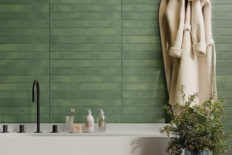 Helles Badezimmer mit grünen horizontalen Wandfliesen, schwarzen Armaturen, weißer Ablage mit Seifenspendern und einem Bademantel.