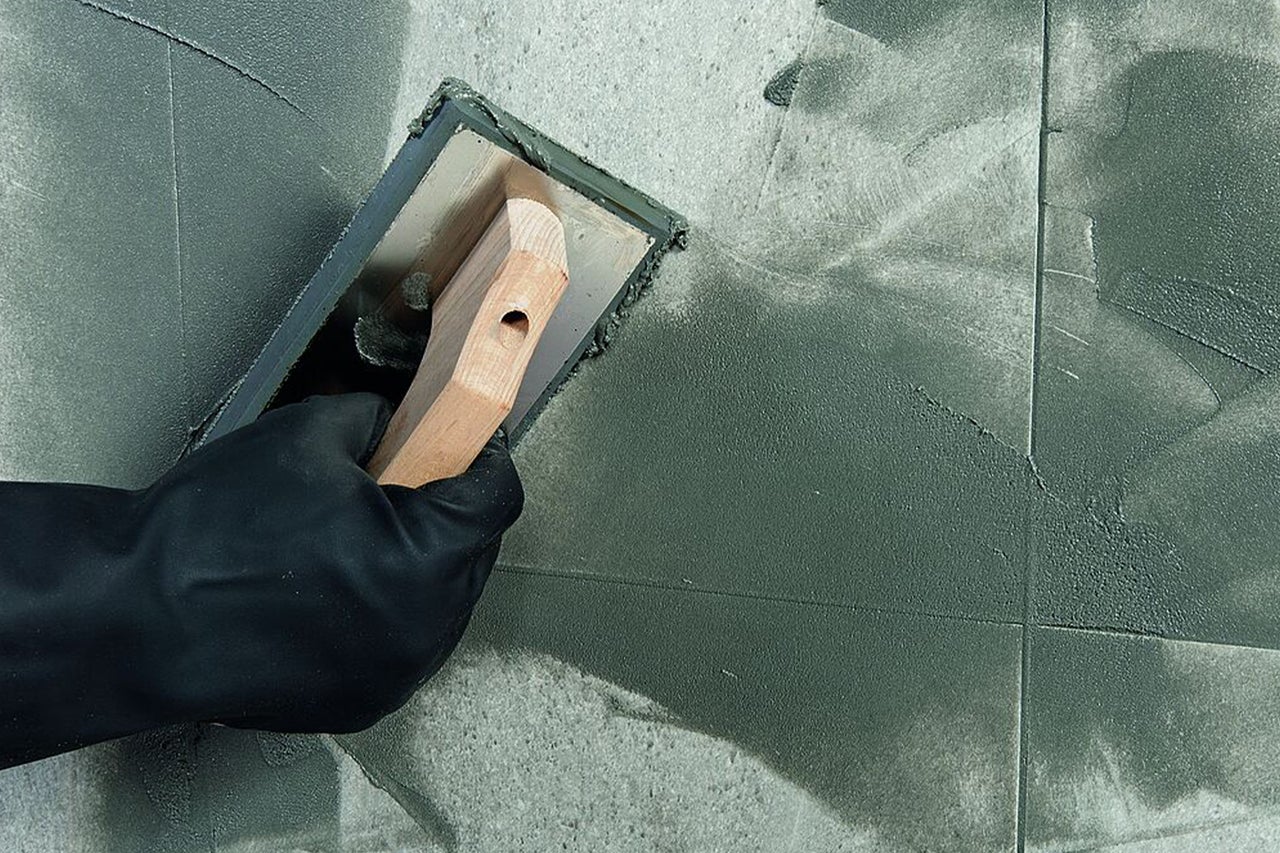 Eine Hand mit einem schwarzen Handschuh trägt mit einem Fugenbrett Fugenmörtel auf Bodenfliesen auf.