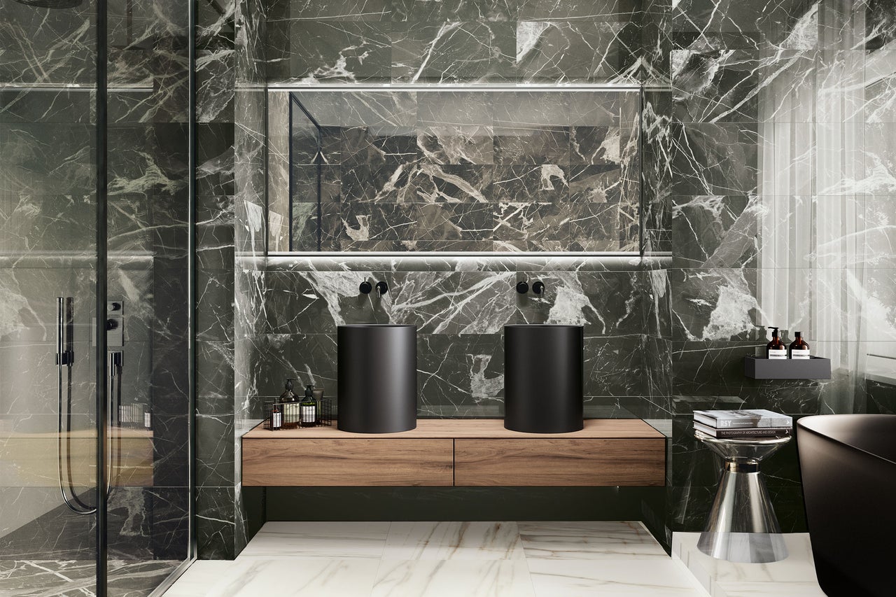 Modernes Badezimmer mit dunklen Marmorfliesen, zwei mattschwarzen Aufsatzwaschbecken auf einem Holzunterschrank und beleuchtetem Spiegel.