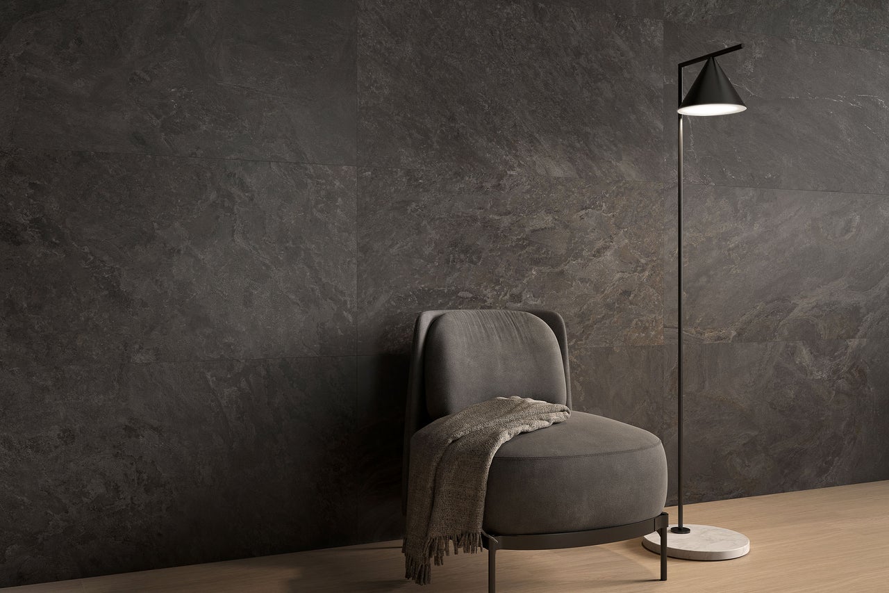 Modernes Wohnzimmer mit großformatigen Wandfliesen in dunkler Natursteinoptik, einem grauen Sessel und einer minimalistischen Stehlampe auf hellem Boden