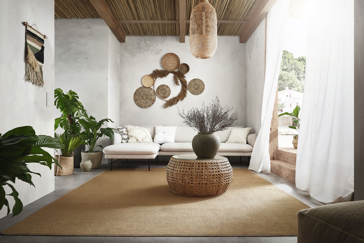 Helles Wohnzimmer im Boho-Stil mit beigem Sofa, rundem Geflecht-Couchtisch, großem Teppich, Zimmerpflanzen und Wanddekoration aus Naturmaterialien.