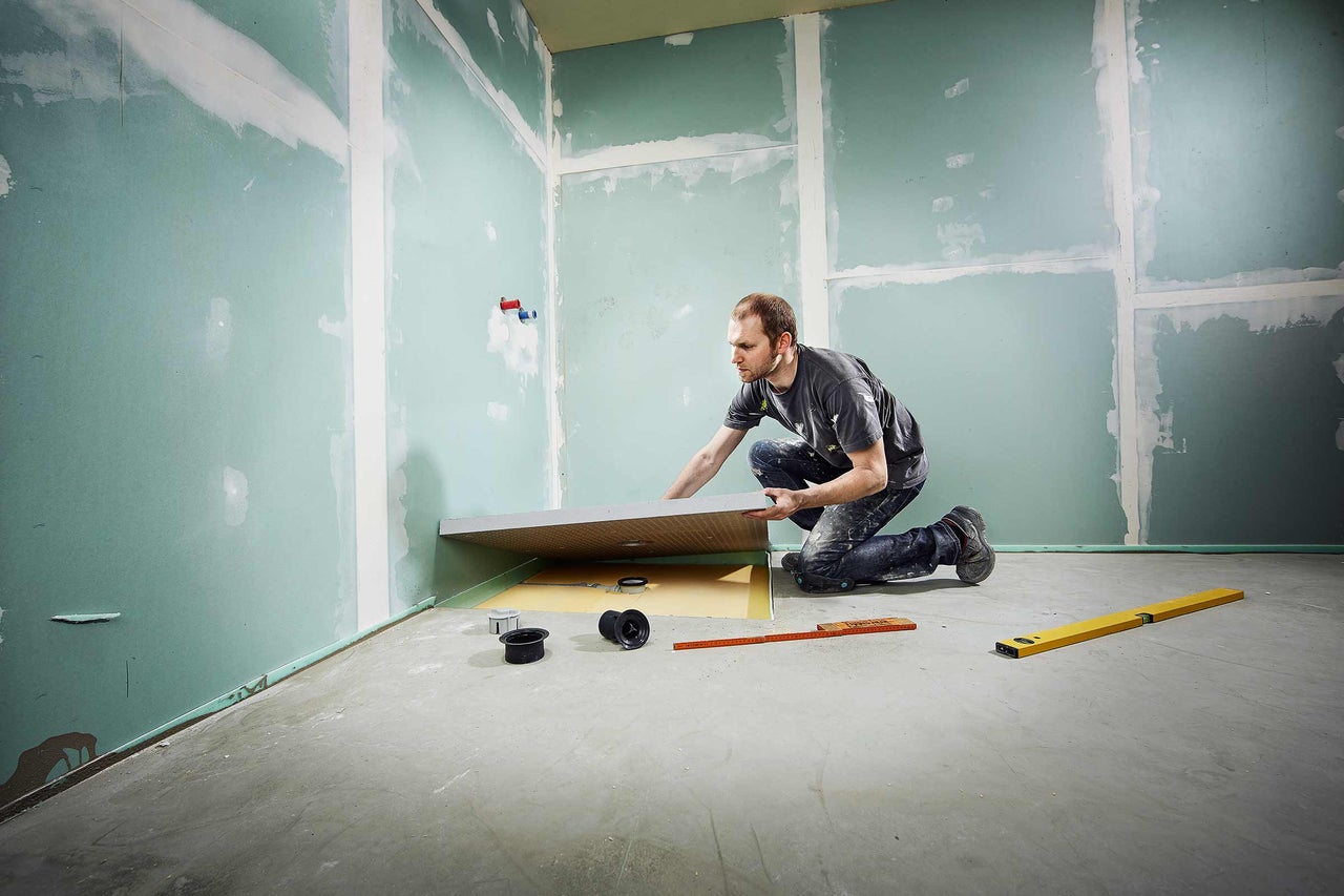 Mann montiert eine Duschtasse in einem Badezimmer mit Trockenbauwänden und Nivellierwerkzeug.