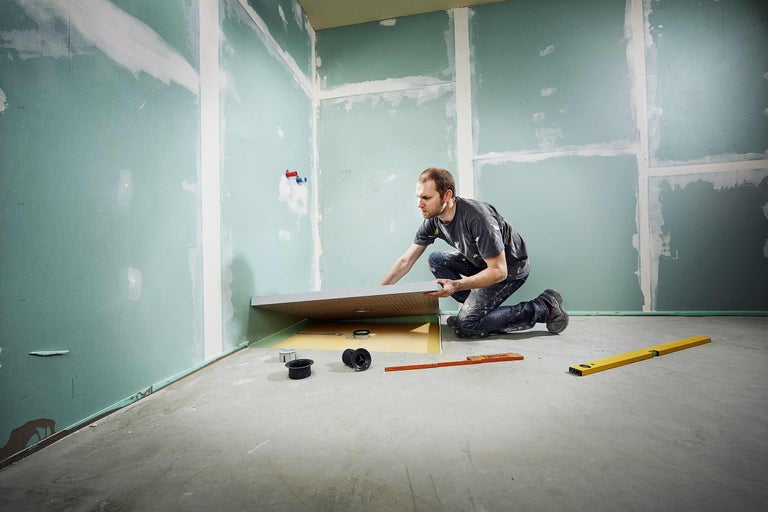 Mann montiert eine Duschtasse in einem Badezimmer mit Trockenbauwänden und Nivellierwerkzeug.