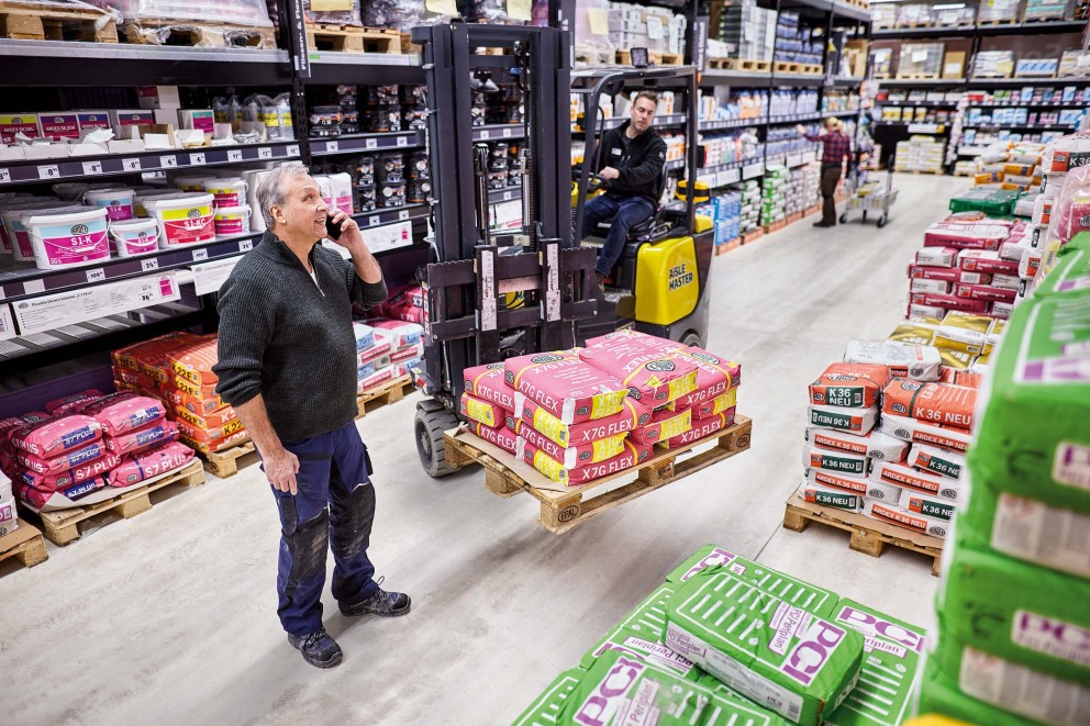 Innenansicht eines Hornbach Baumarktes mit einem Mann, der telefoniert, und einem Gabelstapler, der Fliesenkleber transportiert.